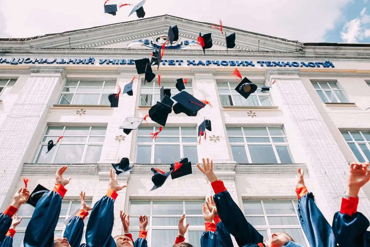 A person in a graduation gown holding a certificate and standing in a graduation ceremony with other students throwing their hats in the air.